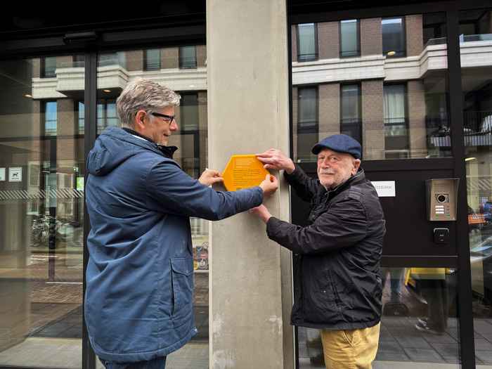 Twee mensen plaatsen een geel herdenkingsbord op de gevel van een gebouw